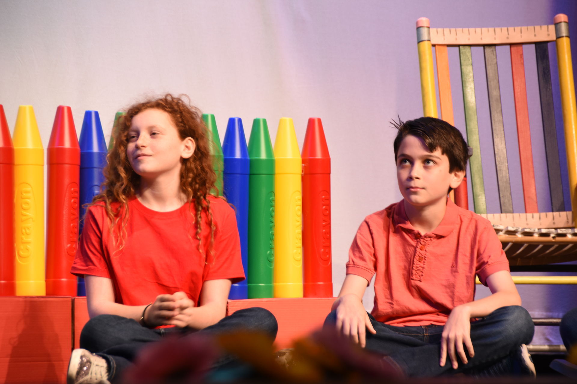 Two children seated in front of a crayon fence and a multicolored chair.
