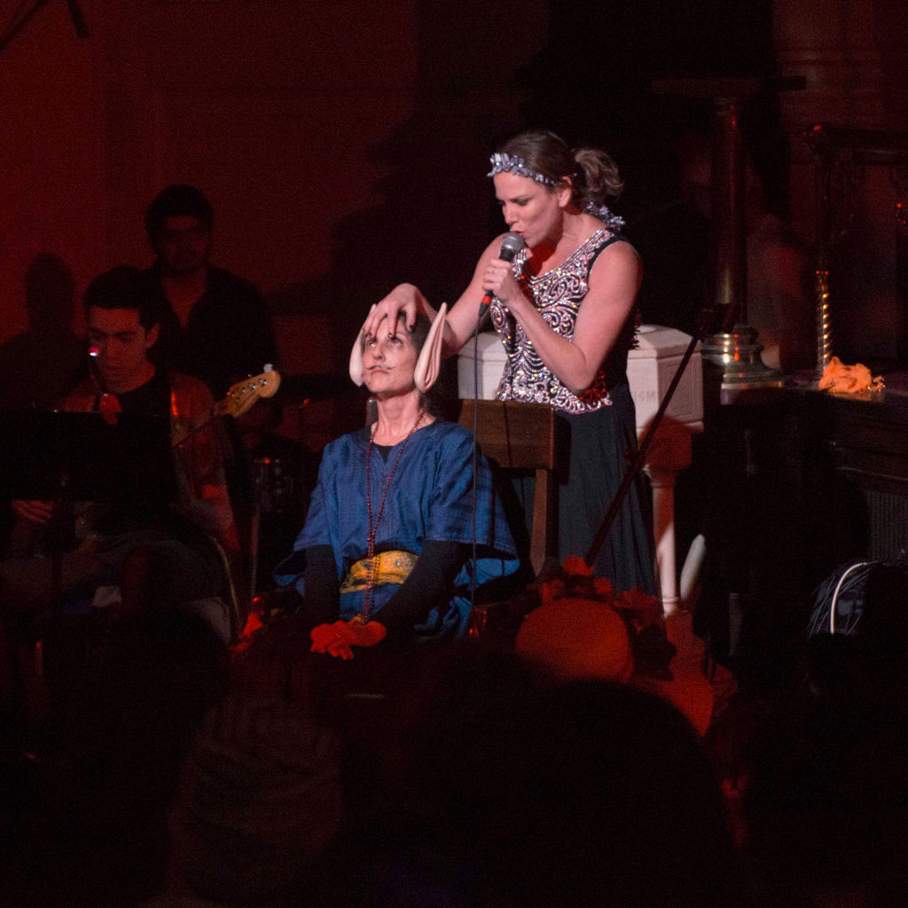 A woman seated in a chair in front of another woman, who is stroking the first woman's hair.