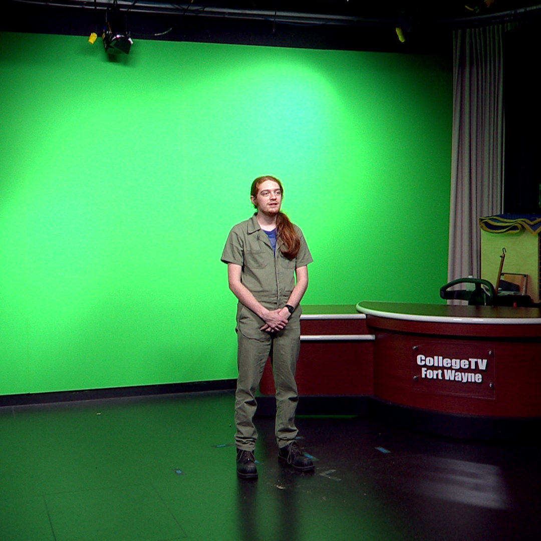Person standing in front of greenscreen, behind them is a desk labeled CollegeTV Fort Wayne.